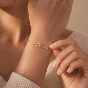 Peut inclure: Un bracelet en or délicat avec le nom "Olivia" en écriture cursive. Le bracelet est porté sur un poignet, mettant en valeur la fine chaîne et le design personnalisé. Le bracelet est un bijou.