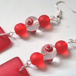 May include: A pair of earrings featuring red rectangular pendants and beaded accents. The earrings have silver-colored fish hook ear wires. The design includes red and clear glass beads, creating a striking contrast.