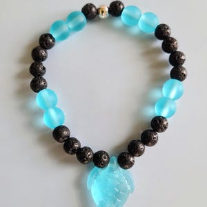 Könnte beinhalten: Ein Armband mit abwechselnd schwarzen Lavastein- und hellblauen Glasperlen. Ein hellblauer Seeschildkröten-Anhänger hängt in der Mitte. Das Armband enthält eine silberfarbene Perle.