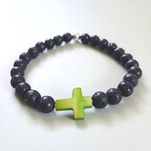 May include: Black lava stone bead bracelet with a bright green cross-shaped charm. The bracelet features a stretch design and a silver-colored clasp. The beads are round and have a textured surface.