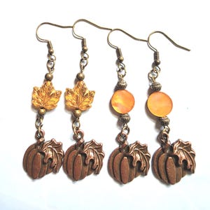 May include: Two pairs of dangle earrings featuring bronze-toned pumpkin charms with attached leaves. One pair includes amber-colored leaf-shaped beads, while the other pair features round orange beads. All earrings have bronze-toned metal hooks.