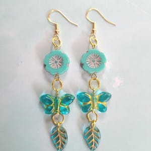May include: A pair of dangle earrings featuring a teal flower, a teal butterfly, and a teal leaf with gold accents. The earrings have gold-colored ear wires and connecting rings. The earrings are displayed against a light blue background.