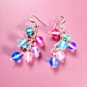 May include: A pair of dangle earrings with silver-tone hooks and chains. Each earring features a cluster of colorful, iridescent glass beads in shades of blue, pink, purple, and green. The earrings are set against a pink background.