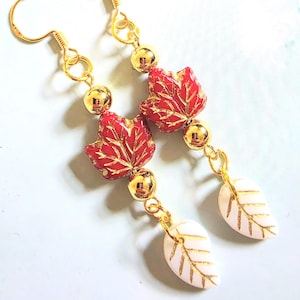 May include: A pair of dangle earrings featuring gold-tone hooks, polished gold-tone spherical beads, red maple leaf-shaped beads with gold veining, and smaller white leaf-shaped beads with gold veining, all connected by gold-tone metal jump rings.