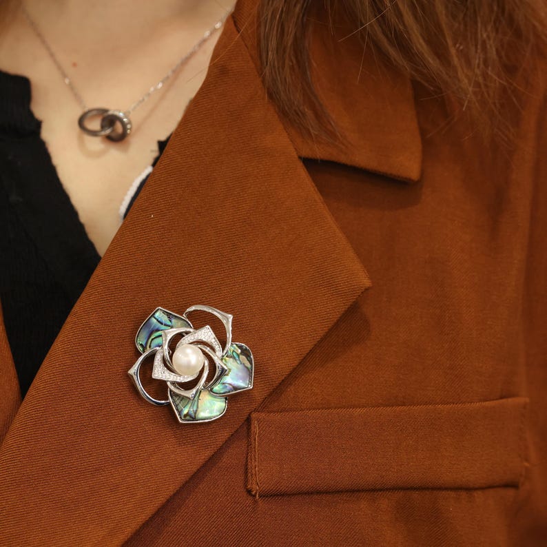 May include: A silver and abalone shell flower brooch with a pearl in the center. The brooch is pinned to a brown jacket.