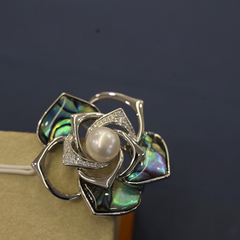 May include: A silver brooch in the shape of a flower with a pearl in the center. The flower petals are made of abalone shell with iridescent green, blue, and purple colors.