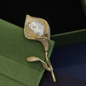May include: A gold-tone brooch in the shape of a calla lily with a large, white baroque pearl in the center. The flower is embellished with clear rhinestones.