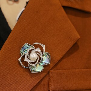 May include: A silver flower brooch with a pearl in the center and abalone shell petals. The brooch is pinned to a brown fabric background.