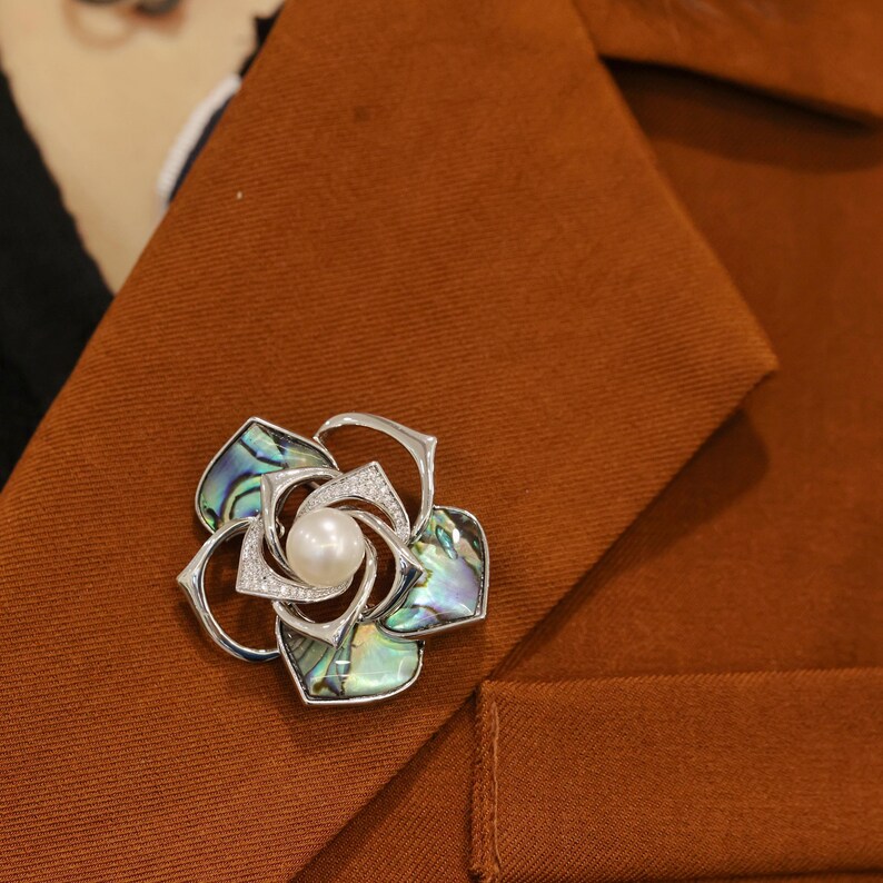 May include: A silver flower brooch with a pearl in the center and abalone shell petals. The brooch is pinned to a brown fabric background.