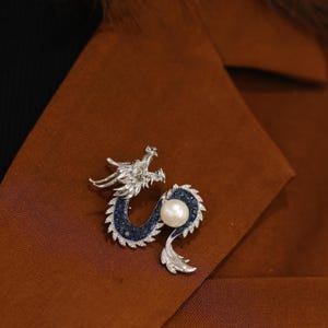 Może przedstawiać: Srebrna broszka w kształcie smoka z perłą i niebieskimi kamieniami szlachetnymi. Ciało smoka otacza perłę, na tle brązowej tkaniny. Unikatowa biżuteria.