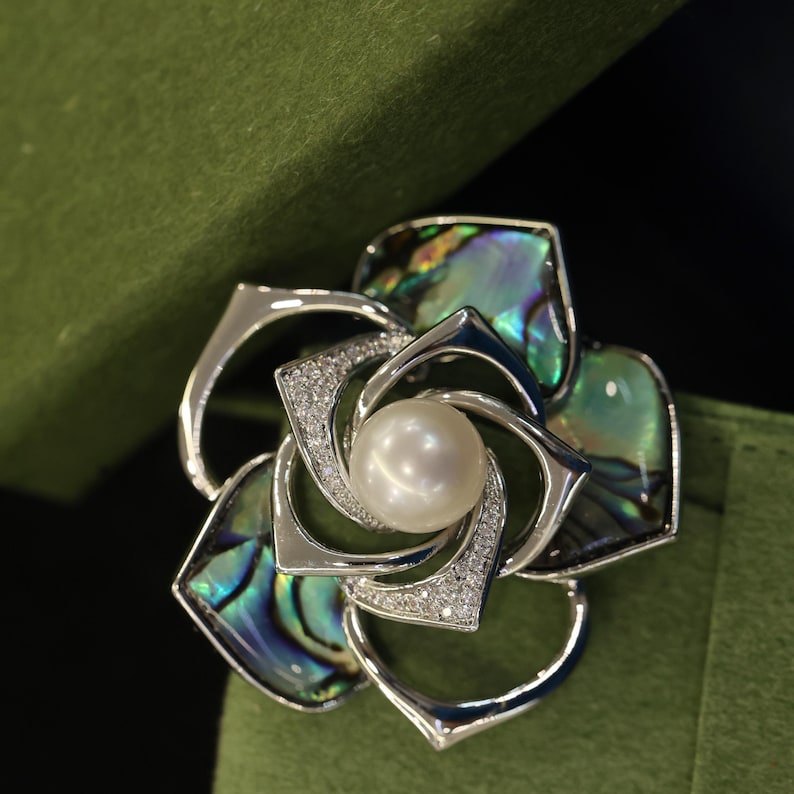 May include: A silver and abalone shell flower brooch with a pearl in the center. The flower has a swirl design and is surrounded by small clear stones.