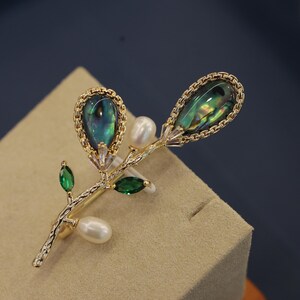 May include: A gold-tone brooch featuring a delicate branch with two green leaves, two faux pearls, and two iridescent green abalone shell accents.