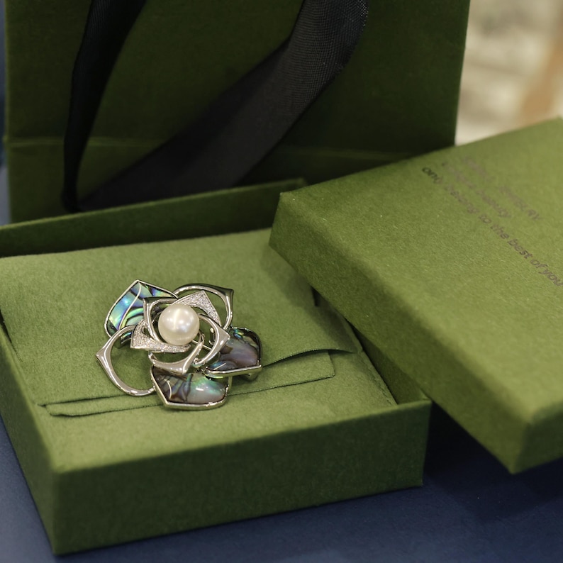 May include: A silver and abalone shell flower brooch with a white pearl in the center. The brooch is displayed in a green velvet box.