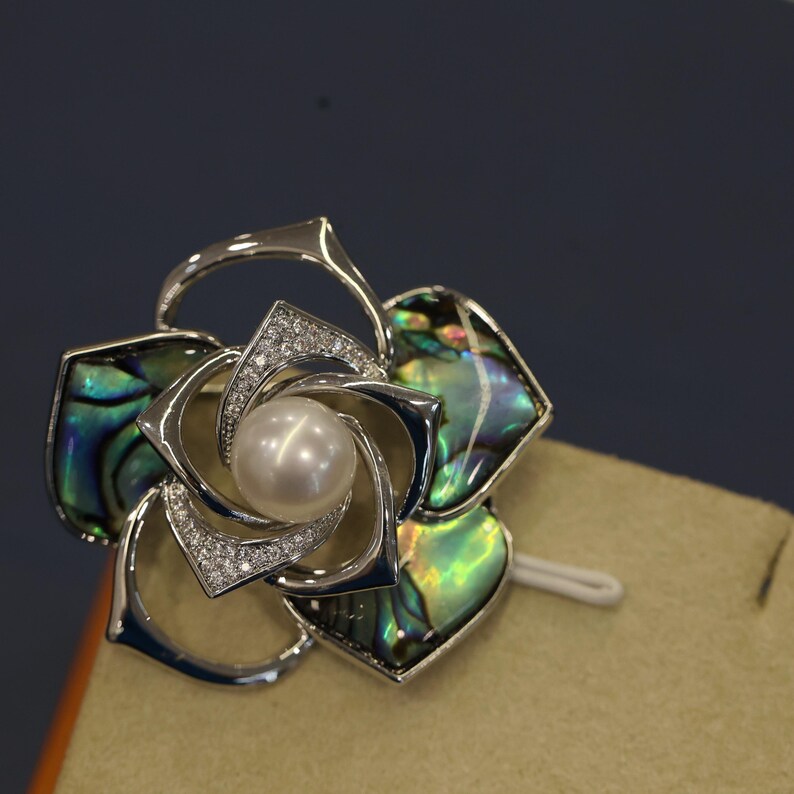 May include: A silver flower brooch with a pearl in the center and abalone shell accents. The flower has a swirling design and is set against a brown background.