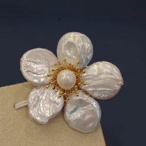 May include: A white pearl flower brooch with a gold center and a single pearl in the middle. The flower has five petals.