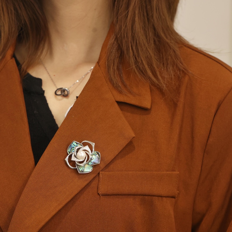 May include: A silver brooch with a pearl in the center and a flower design made of abalone shell. The brooch is pinned to a brown jacket.