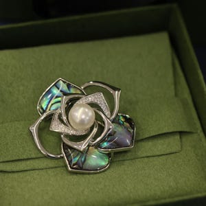May include: A silver brooch in the shape of a flower with a pearl in the center. The flower is made of abalone shell with iridescent green, blue, and purple colors.