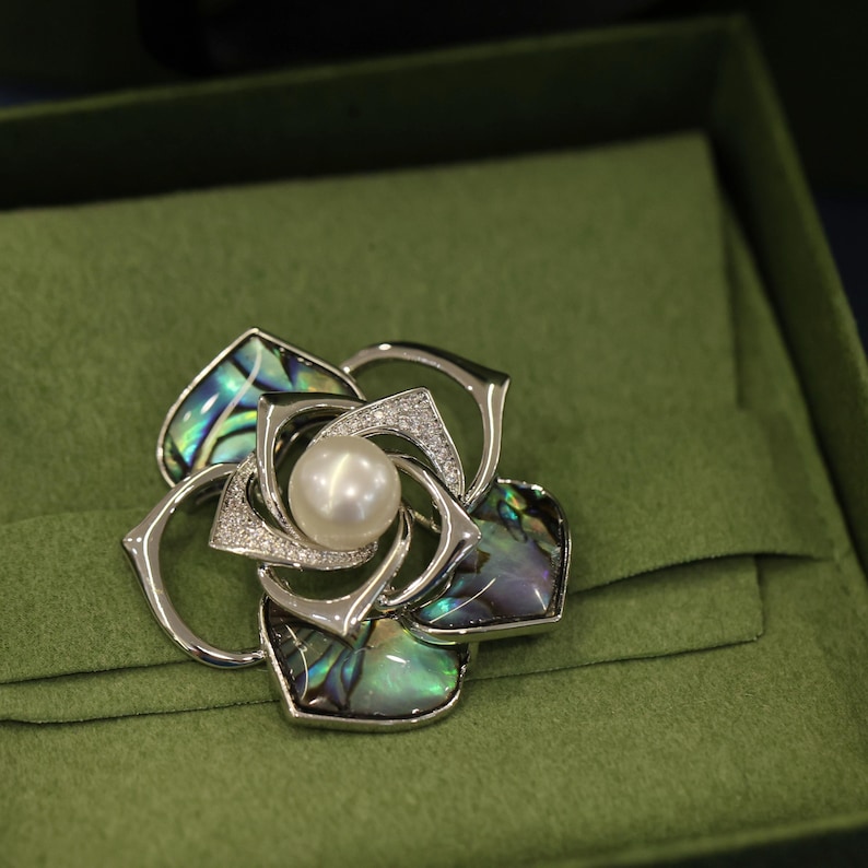 May include: A silver brooch in the shape of a flower with a pearl in the center. The flower is made of abalone shell with iridescent green, blue, and purple colors.