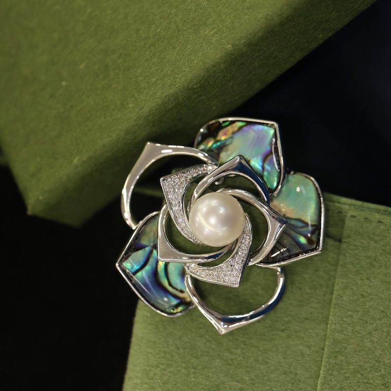 May include: A silver brooch in the shape of a flower with a pearl in the center. The flower petals are made of iridescent abalone shell.