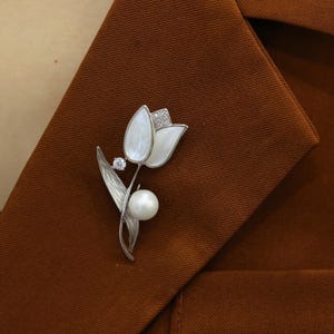 Puede incluir: Un broche de flor de tulipán blanco con una perla y diamantes de imitación. El broche es de color plateado y tiene un diseño delicado.