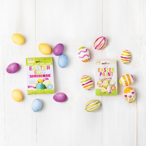 May include: An overhead shot of Easter eggs and coloring kits on a white wooden surface. The eggs are painted in various colors and patterns. Two boxes read "Easter Egg Coloring Kit" and "Easter Paint".