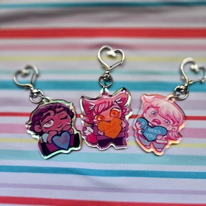 May include: Three anime-style keychains with heart-shaped clasps. Each keychain features a different character holding a heart-shaped candy with text. The characters are in shades of pink, purple, and blue.