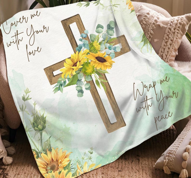 Może przedstawiać: Biały koc z drewnianym krzyżem ozdobionym ż&oacute;łtymi słonecznikami i zielonymi liśćmi. Na kocu nadrukowano frazy "Cover me with Your love" i "Wrap me with Your peace". Koc jest narzucony na krzesło.