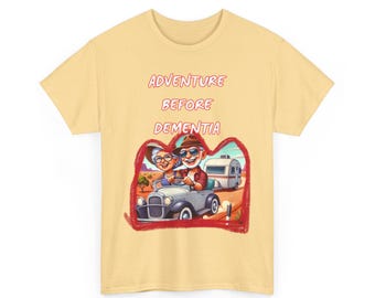 Op de afbeelding: Een geel T-shirt met de tekst "Adventure Before Dementia" in rood. Het shirt heeft een cartoonillustratie van een ouder echtpaar dat een oldtimer rijdt met een caravan erachter.