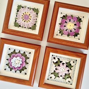 May include: Four framed crochet flower art pieces. Each square frame features a different colorful floral design, including pink, purple, and white flowers with green leaves, set against a white background. The frames are made of brown wood.