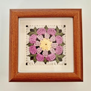 May include: A framed square crochet piece featuring a floral design. The flower has a yellow center, surrounded by pink petals and green leaves, set against a white background. The frame is made of brown wood.