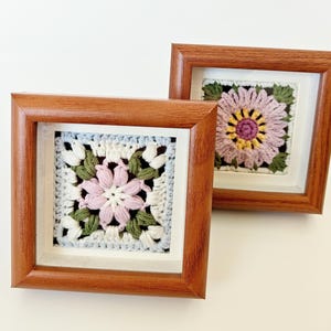 May include: Two square framed crochet art pieces. Each features a detailed flower design in shades of pink, white, green, and yellow. The frames are made of brown wood and are displayed against a white background. The crochet work is intricate and colorful.