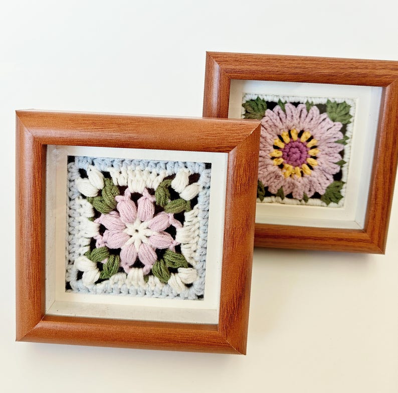 May include: Two square framed crochet art pieces. Each features a detailed flower design in shades of pink, white, green, and yellow. The frames are made of brown wood and are displayed against a white background. The crochet work is intricate and colorful.
