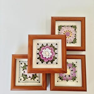 May include: Four framed crochet art pieces. Each square frame is made of wood and contains a crocheted flower design in shades of pink, white, and green. The crochet work is set against a white background.