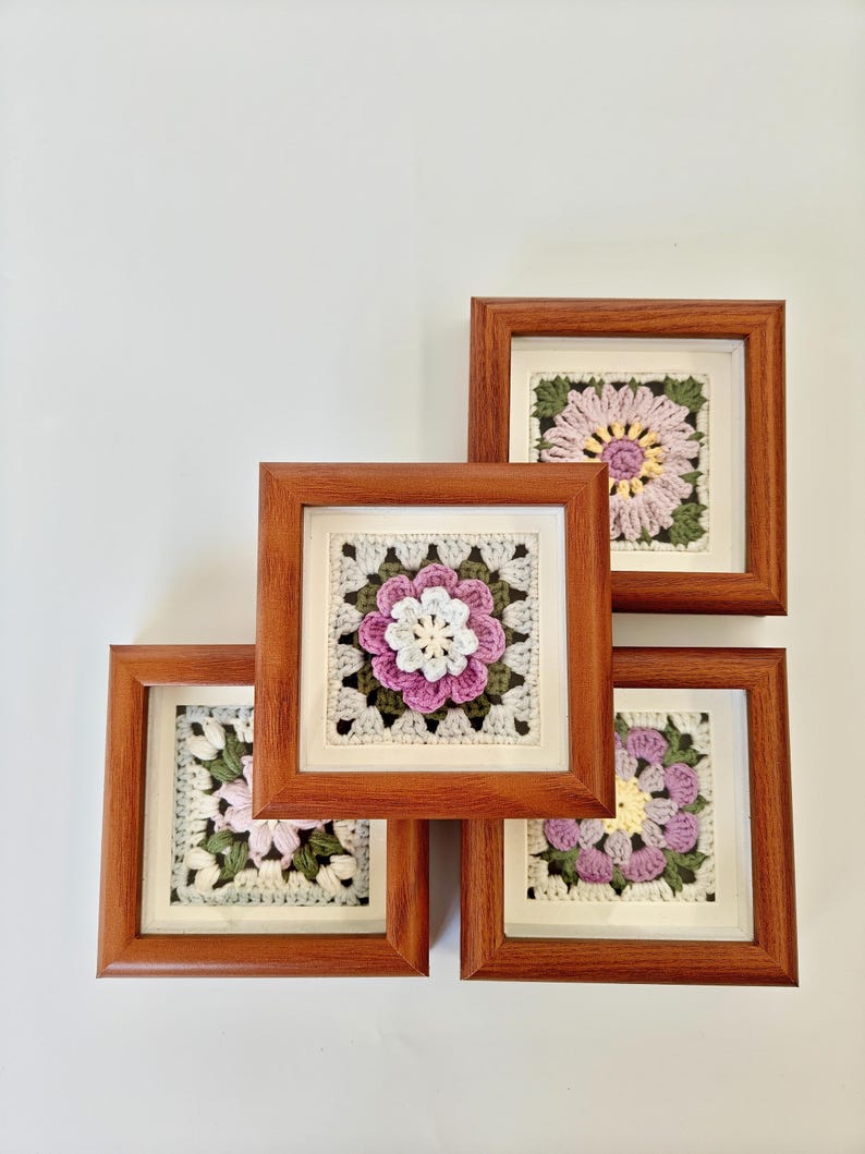 May include: Four framed crochet art pieces. Each square frame is made of wood and contains a crocheted flower design in shades of pink, white, and green. The crochet work is set against a white background.