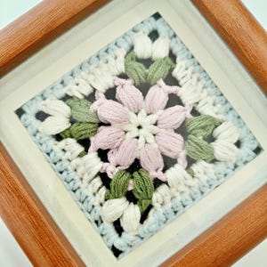 May include: A framed, square crochet art piece. The artwork features a central flower design in pink, surrounded by green and white petals, set against a light blue and white background. The frame is made of wood.