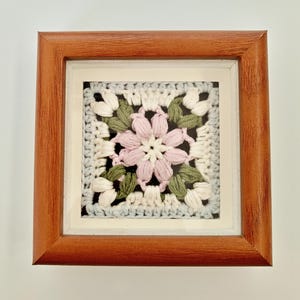 May include: Square framed crochet art featuring a central pink flower with white and green leaves. The crochet piece is set against a light blue and white background, framed in a brown wooden frame.