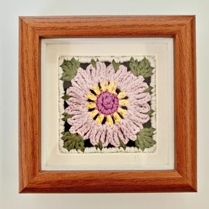 May include: A framed, square crochet art piece featuring a large, stylized flower. The flower has pink petals, a yellow center, and a purple center. Green leaves surround the flower, set against a black background. The frame is made of brown wood.