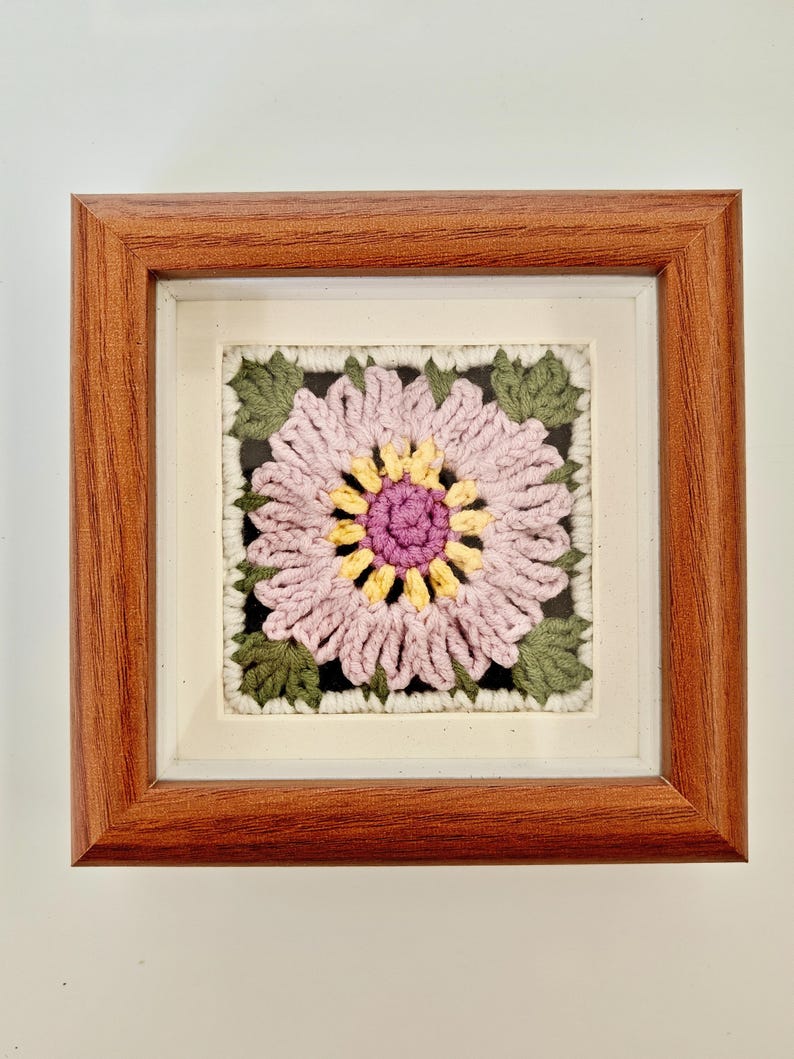 May include: A framed, square crochet art piece featuring a large, stylized flower. The flower has pink petals, a yellow center, and a purple center. Green leaves surround the flower, set against a black background. The frame is made of brown wood.