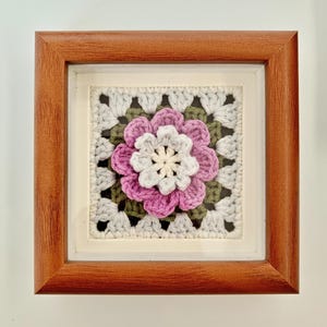 May include: A framed square crochet art piece featuring a central flower design. The flower has white, pink, and purple petals, surrounded by green and gray crochet work. The frame is made of brown wood.