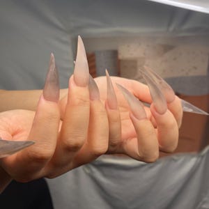 Puede incluir: Una mano con uñas acrílicas largas y transparentes. Las uñas son puntiagudas y se extienden más allá de las yemas de los dedos. Las uñas tienen un aspecto translúcido, con un sutil degradado de color.