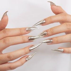 Puede incluir: Uñas de estilo stiletto en plata metálica. Las uñas son largas, puntiagudas y tienen un acabado reflectante, similar al cromo. La manicura se muestra sobre un fondo blanco liso.