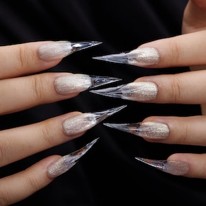 May include: Close-up of a set of long, clear acrylic nails with a sharp, pointed shape. The nails are applied over a shimmering, light-colored nail polish. The background is a solid black, highlighting the nail design.