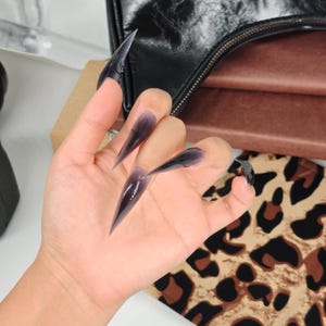 May include: A hand with long, pointed, black and purple ombre acrylic nails. The nails are sharply tapered and glossy. The background includes a black purse and a leopard print pattern.