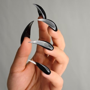 Könnte beinhalten: Künstliche Krallennägel in Schwarz und Silber. Die Nägel sind lang, gebogen und glänzend, mit schwarzer Basis und silbernen Spitzen. Die Nägel werden vor einem grauen Hintergrund gezeigt.