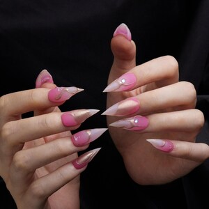 Puede incluir: Uñas acrílicas largas y puntiagudas. Pintadas con un degradado de rosa y transparente, con detalles dorados y pequeños brillantes. Un accesorio de moda para las manos.