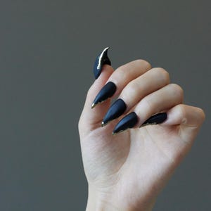 以下が含まれることがあります： ゴールドのアクセントが付いた、長いマットブラックのスティレットネイルを披露する手。 爪は尖っており、先端に金のトリムが付いています。 1 つの爪には金のストライプがあります。 背景は無地のグレーです。