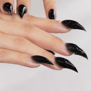 Op de afbeelding: Close-up van een hand met lange, zwarte acrylnagels. De nagels zijn puntig en glanzend, met een gradiënteffect dat overgaat van een lichtere basis naar een diepzwart aan de uiteinden. De nagels zijn klauwachtig.