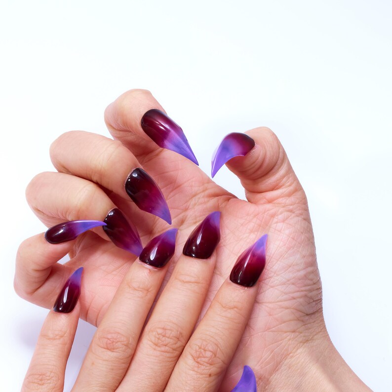 May include: A hand model wearing long, pointed, fake nails with a gradient of dark purple to light purple.