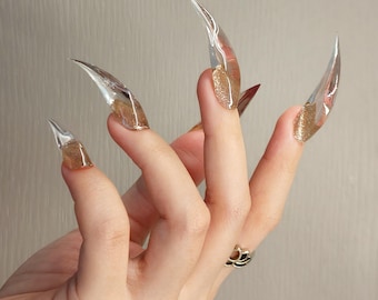 May include: A set of long, stiletto-shaped artificial nails with a clear, transparent finish. Each nail features a decorative gold glitter accent concentrated at the base, creating a gradient effect that extends toward the pointed, translucent tips.