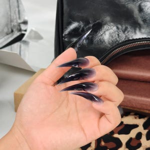 May include: A hand displaying long, pointed, black and purple ombre acrylic nails. The nails have a glossy finish and a sharp, stiletto shape. The nails are the focal point, with a blurred background.